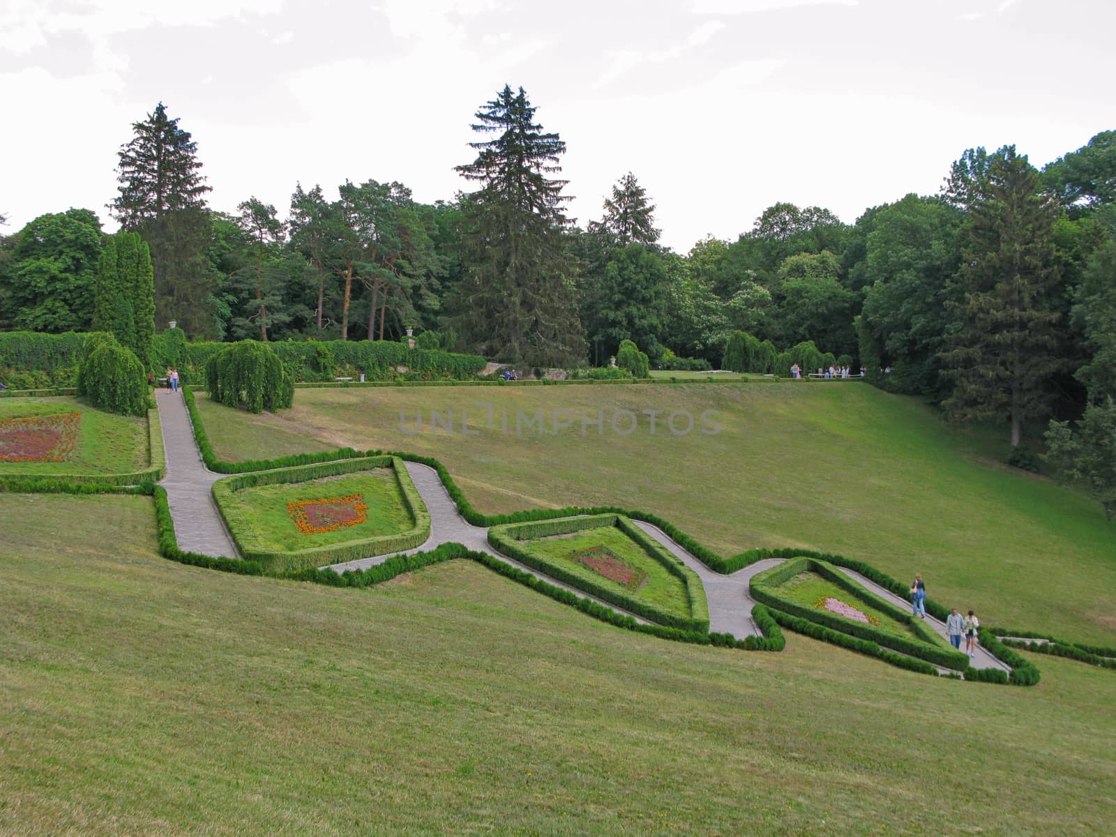 A beautiful landscape of a green lawn in a park on a slope with paths going between figured flower beds with people walking by them.