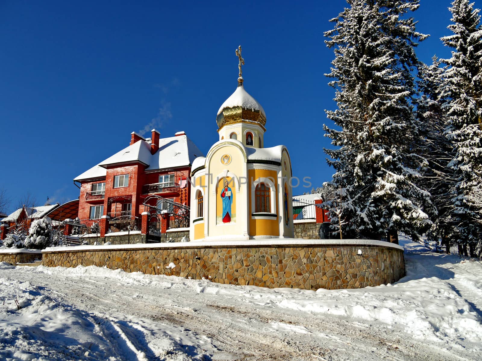 Chapel with the face of the saint on the wall and a dome sprinkled with snow. Standing near a large brick private house against the background of a cloudless winter sky.