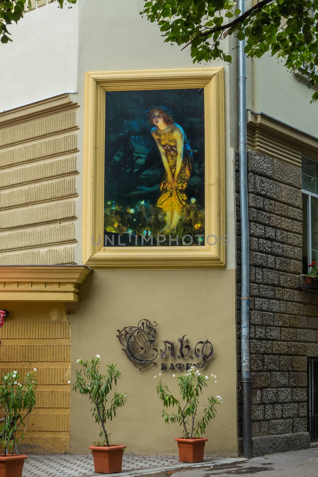 A large picture in the frame with a painted girl on the wall of the house with an inscription cafe under it. And standing flowers in pots on the sidewalk near the house.