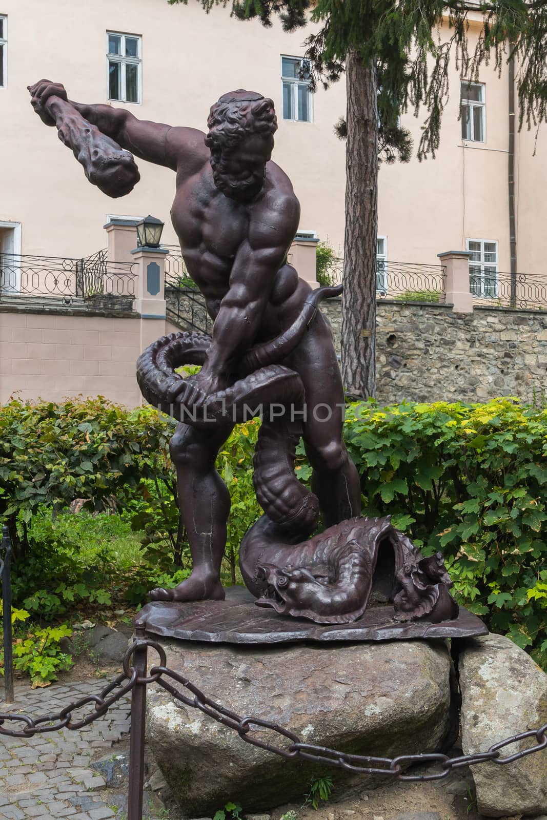 Sculpture depicting a man with a truncheon of a conquering serpent near his feet. Standing on the stones behind the fence with a chain near the green bushes in front of the house. by Adamchuk