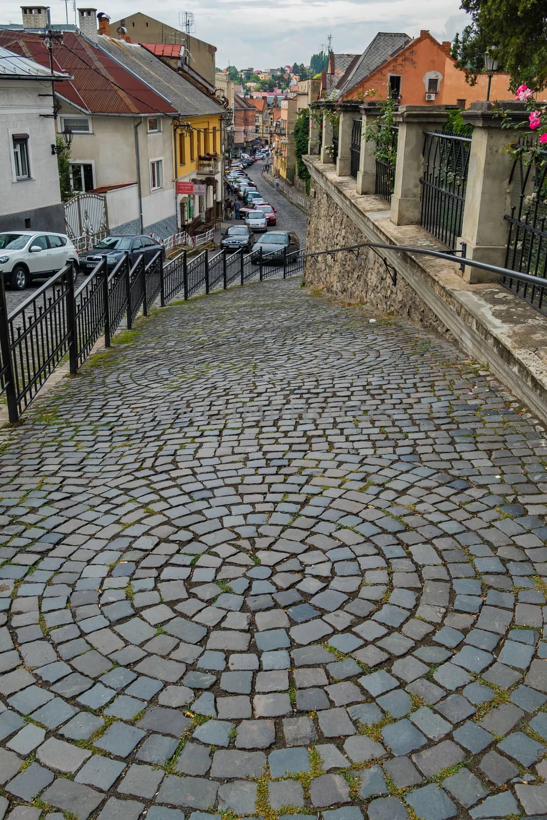 Descent on a city street laid out of tiles with patterns in the form of a circle going down. With nice houses and cars parked on one side. by Adamchuk