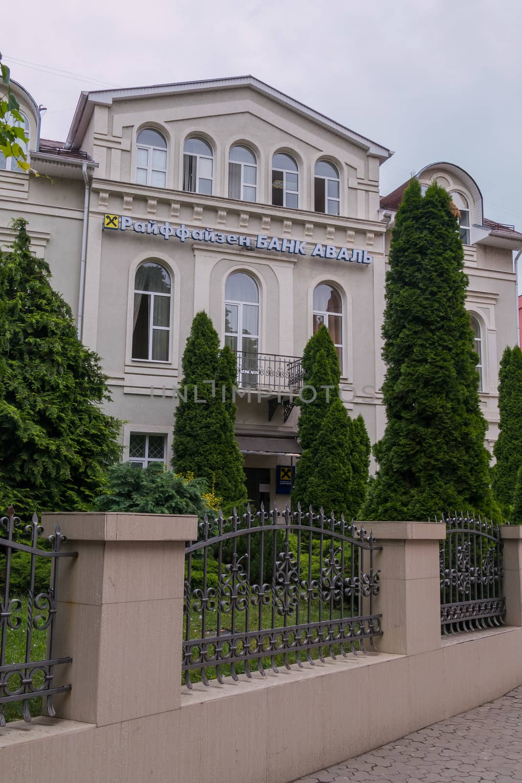The facade of the building with the letters of the name of the organization and the emblem is located behind the unequal fence with bars and green bushes and trees growing side by side. by Adamchuk