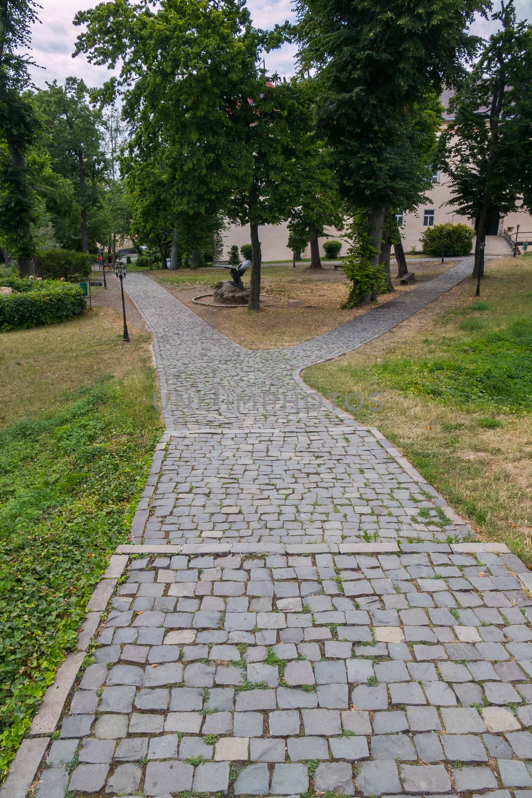 Stepped path in the park running between green trees with standing sculptures on the side of it and leading to the building in the distance.