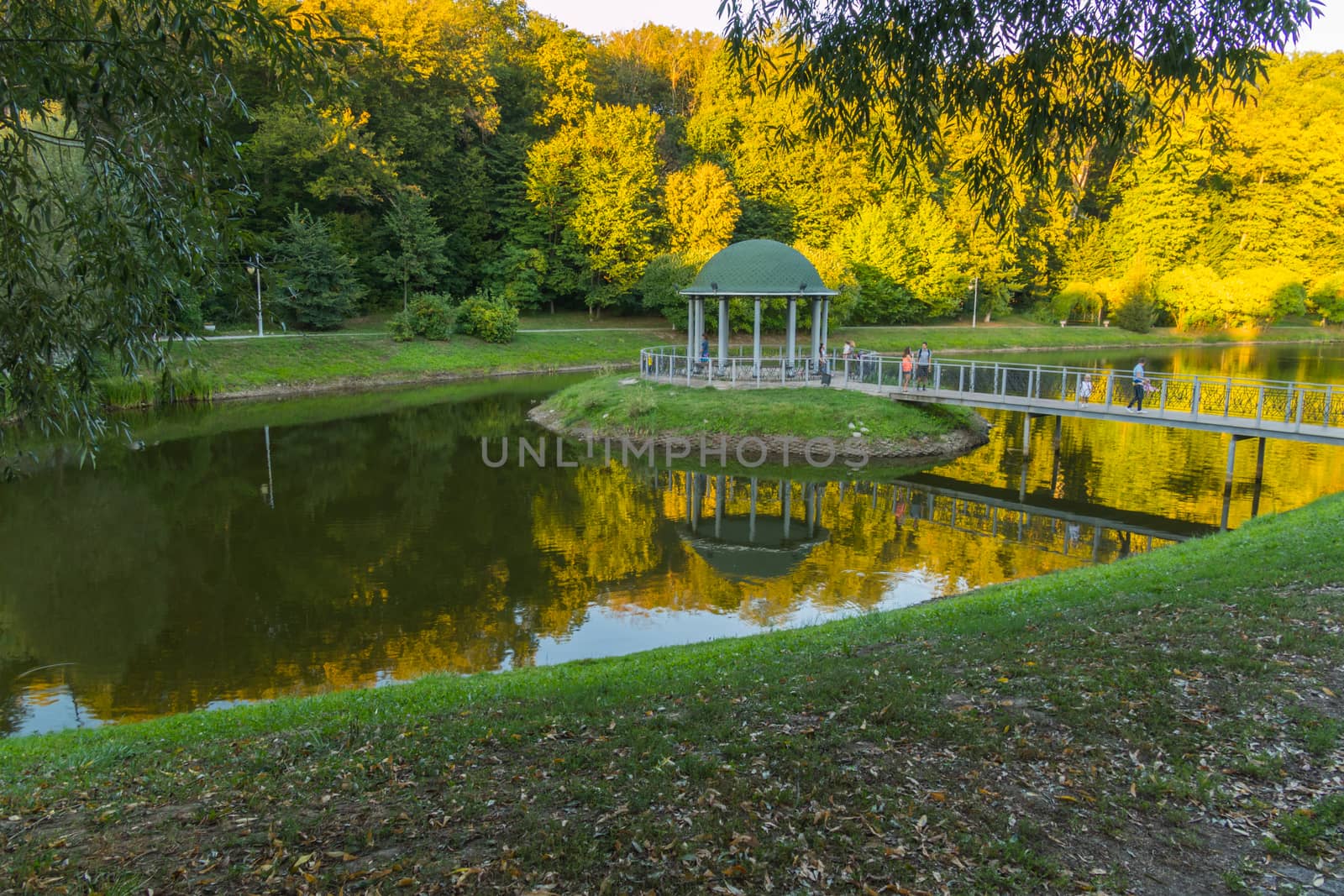 A small island with a decorative alcove with a green roof and a bridge to it against the background of a transparent lake