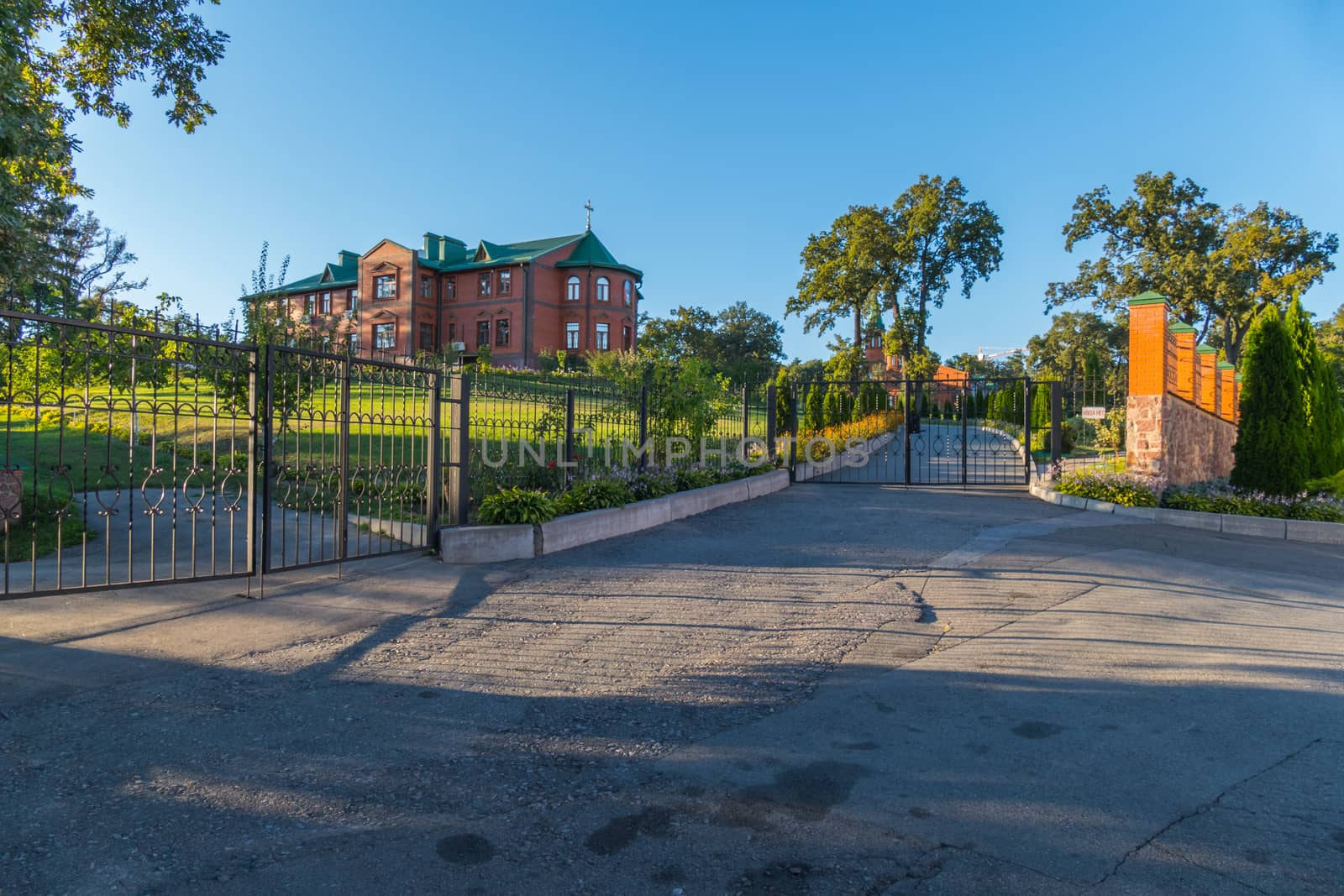 An asphalted playground in front of the gate leading to a beautiful two-story brick house standing on a hill with a beautiful garden in the yard.