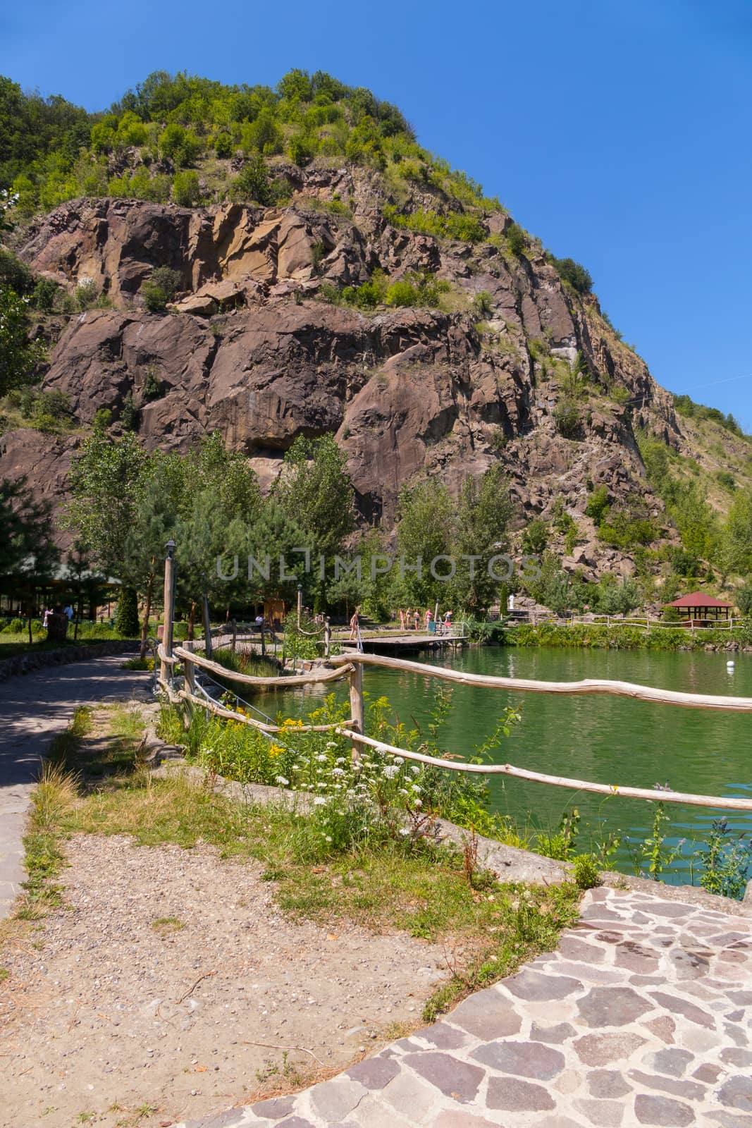 Lake with a gazebo on the shore at the foot of a tall rock on a blue clear sky background