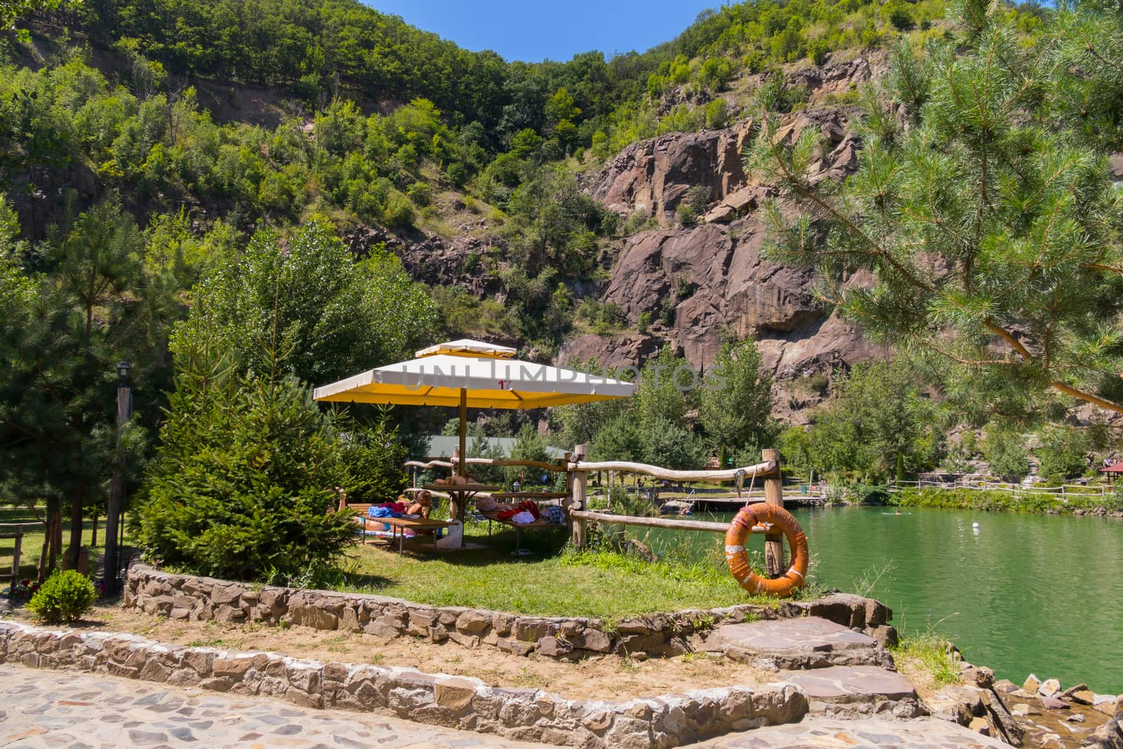 A sun umbrella standing on the shore of a pond near a wooden fence made of poles located at the foot of a stony slope.