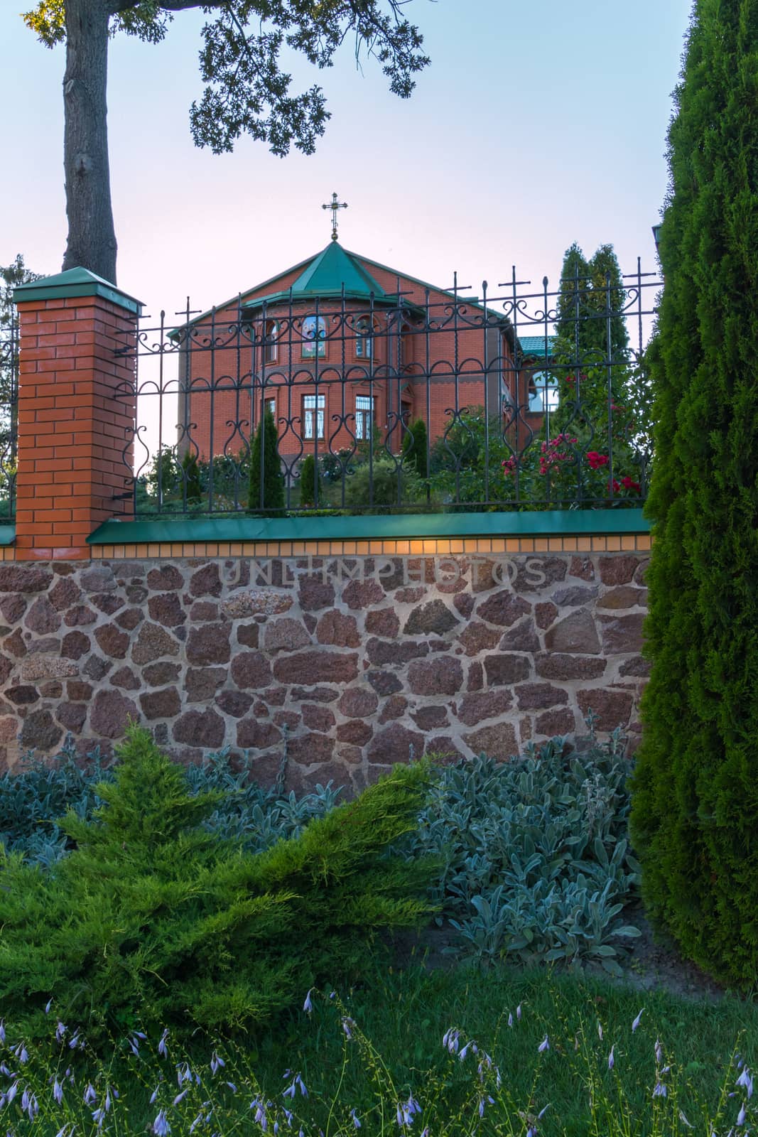 Church of red brick with a green roof behind a forged black high fence by Adamchuk