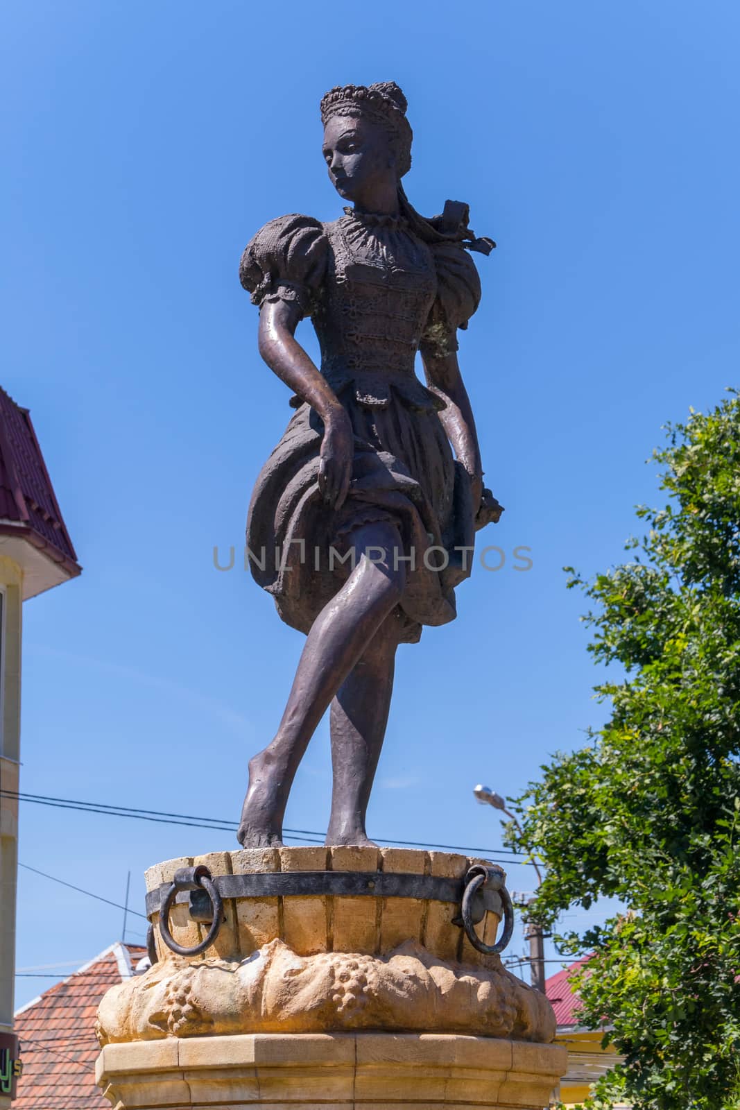 Sculpture of a girl standing on a wooden barrel standing on a pedestal. Against the background of a clear sky without clouds and foliage of trees.