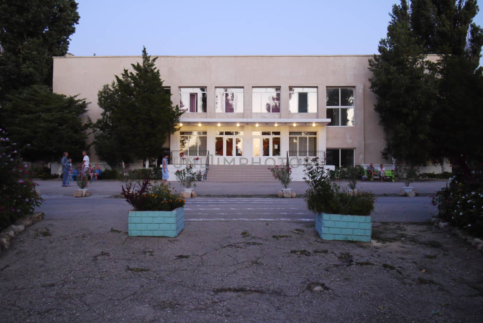 The entrance to the building is illuminated by bulbs under a visor with cracked asphalt near the entrance and flowers in square flowerpots.