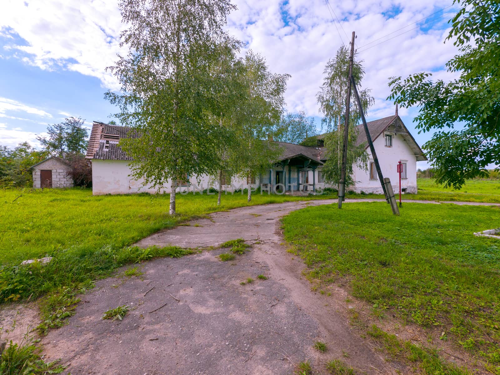 Asphalt cracked road overgrown with grass leading to an abandoned building in the countryside with windows clogged up. by Adamchuk