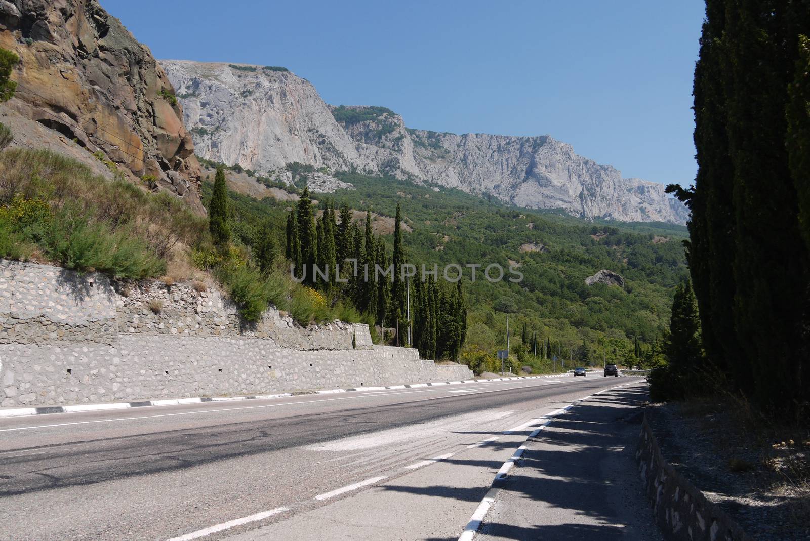 A large motorway on the background of high rocky mountains and green cypresses by Adamchuk