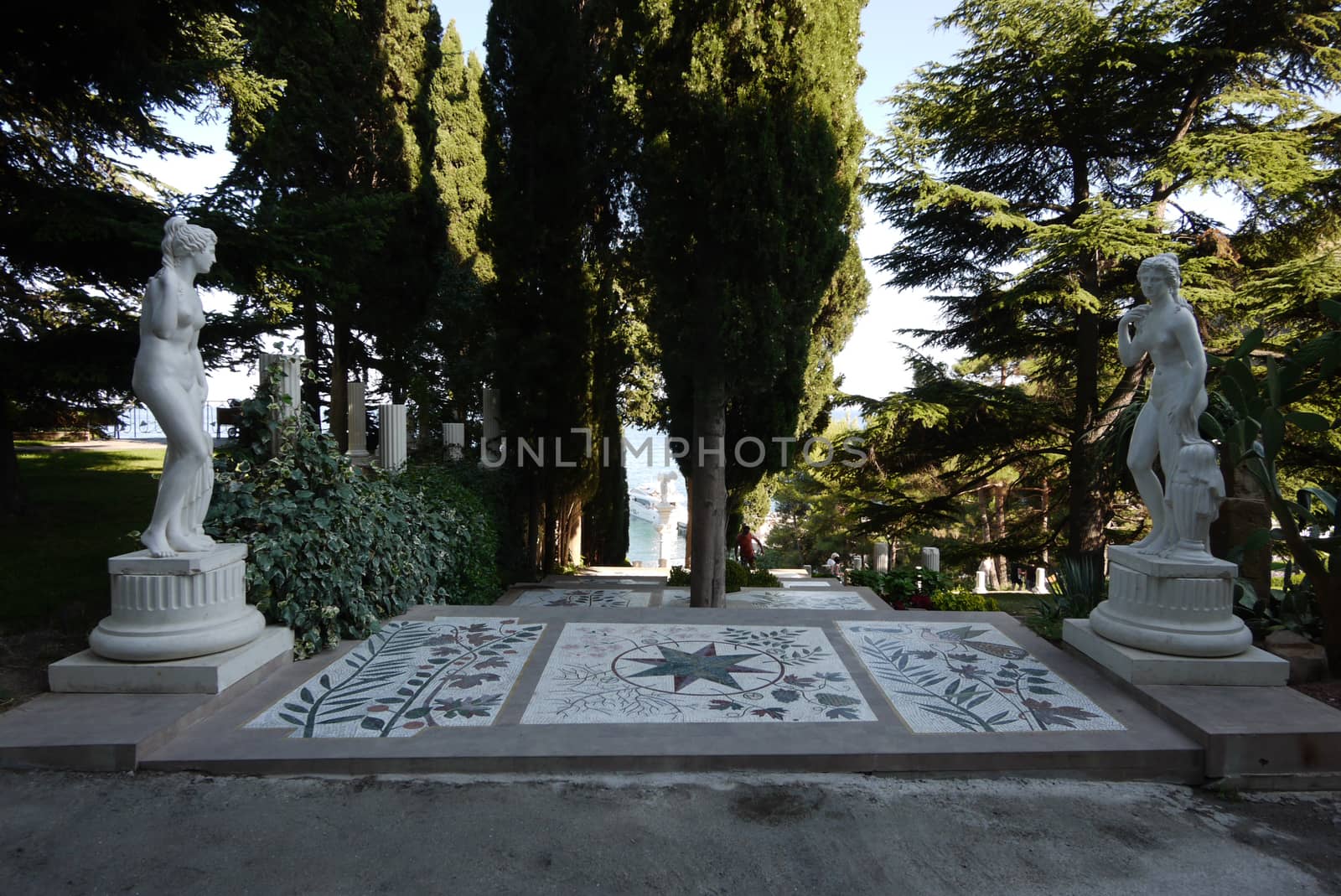 White sculptures in the park are standing from different sides of the path from the plates with a pattern separated by growing tall trees.