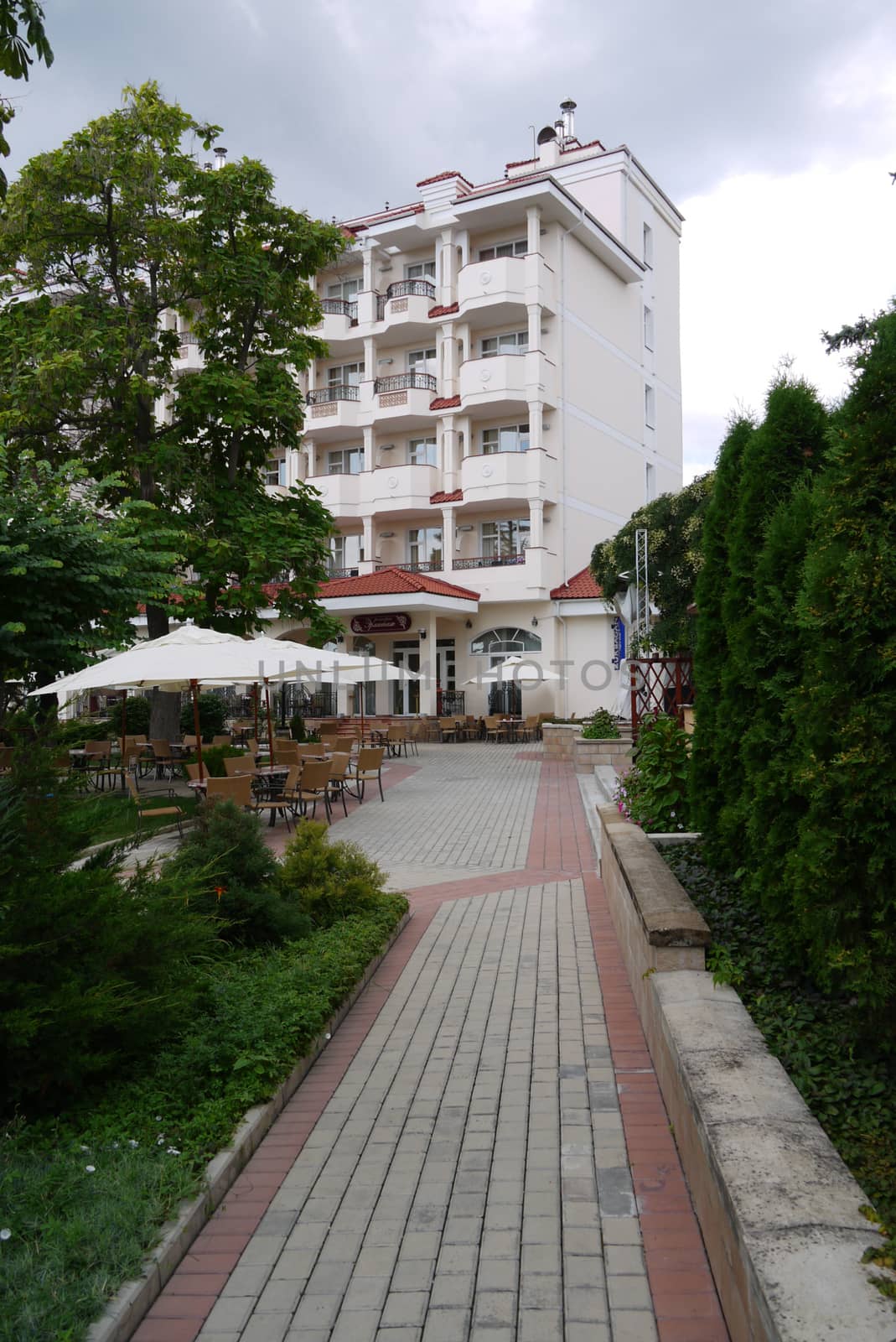 The path running between green bushes and lawn leads to the hotel with balconies with tables and chairs standing next to it for tourists on the street.