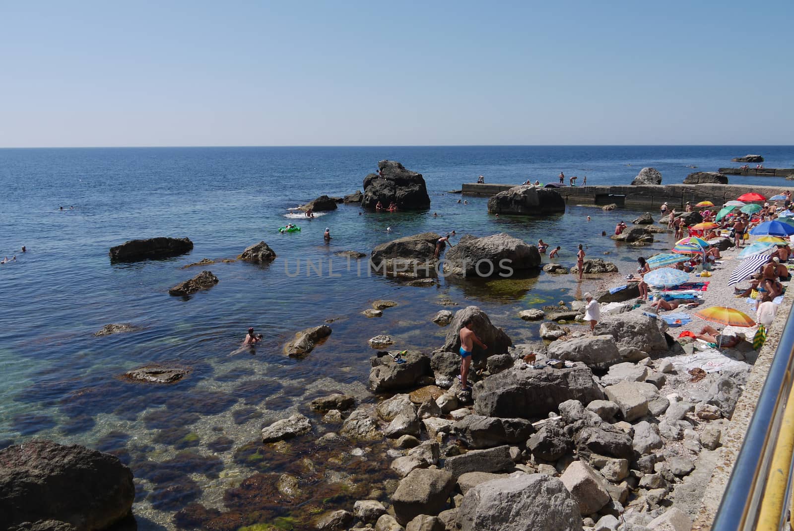 Tourists, resting on the beach next to the large stone boulders, going far into the sea