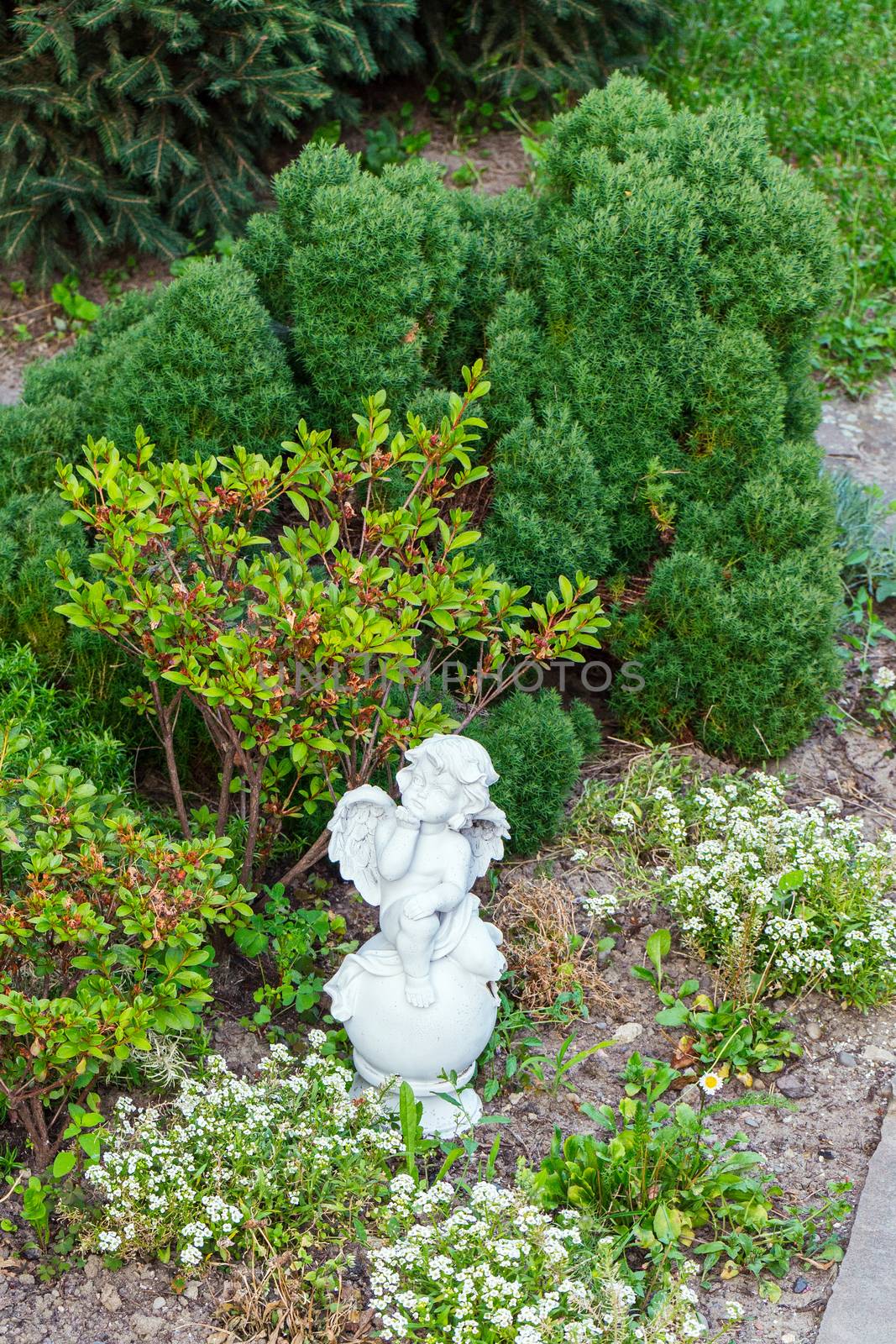 Sculpture of a small angel with a chin resting in the park near green bushes and flowers.