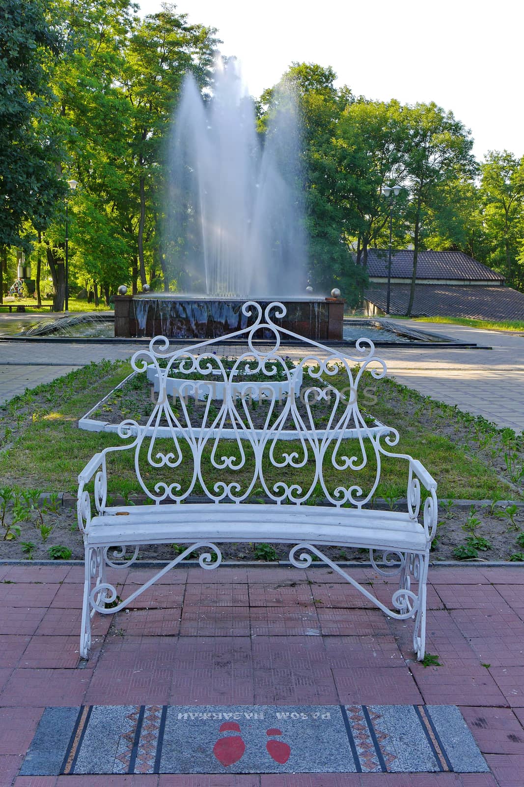 Elegant white bench with a curved back standing in a park near a green lawn in front of a beautiful fountain with clear water jets.
