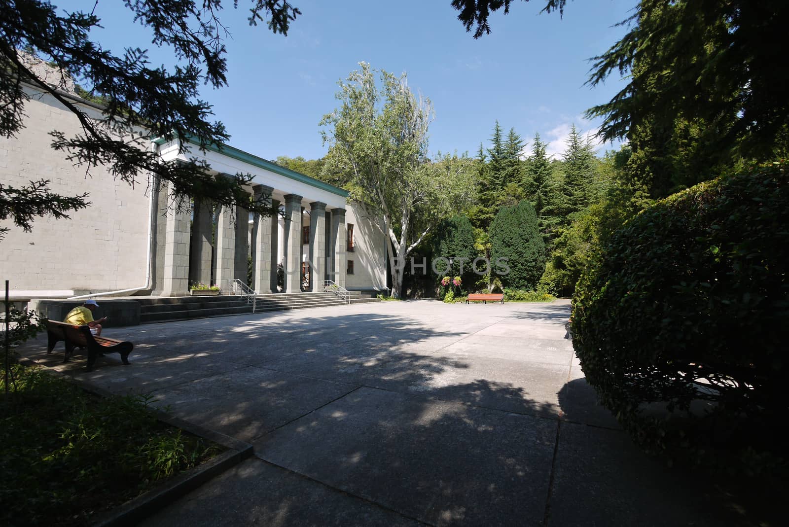 A person resting on a bench in the shade of a tree standing on a platform in front of the park entrance with tall columns.