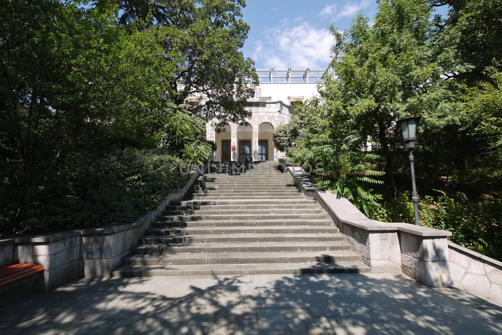 Wide stone steps with three arches at the entrance of the building against the backdrop of green trees and a blue sky by Adamchuk