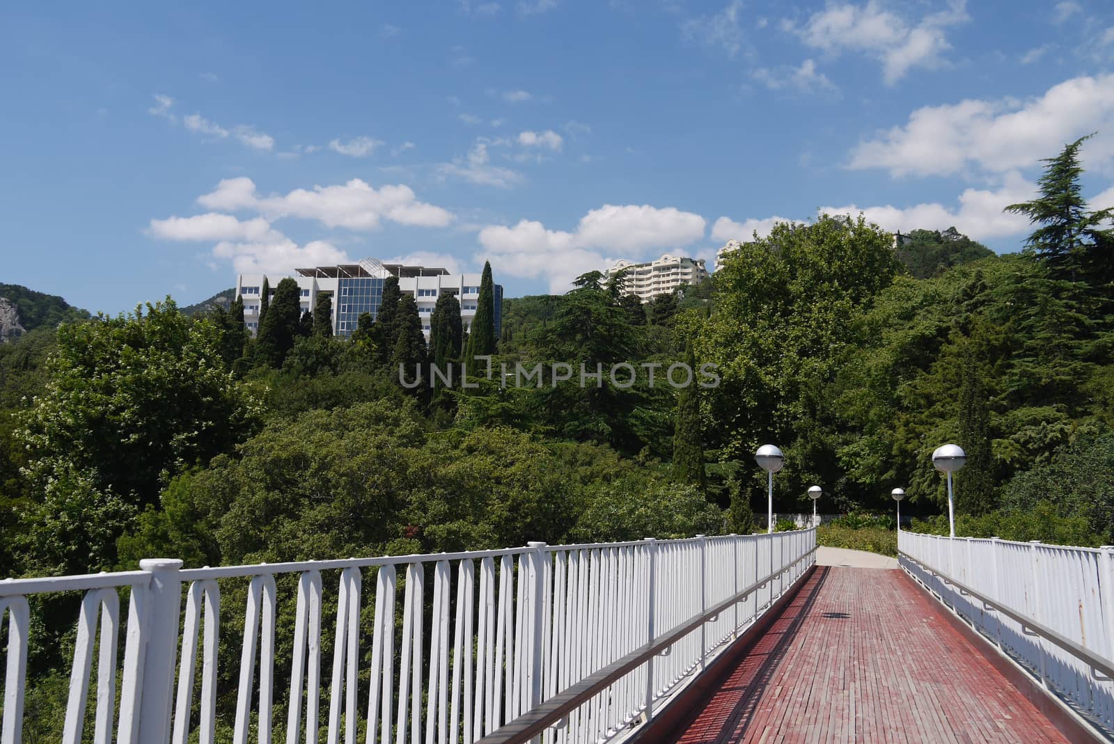 A small bridge with white railings leading to beautiful spa buildings in the shade of green mountain trees by Adamchuk