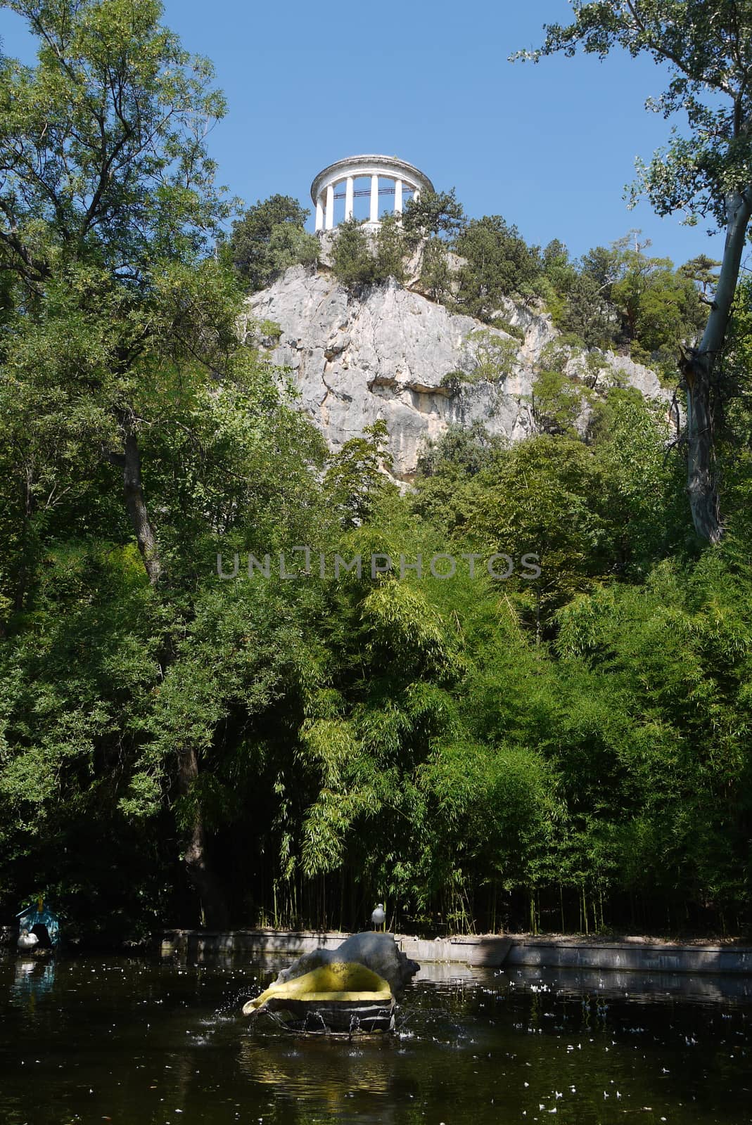 The arbor on top of a rocky cliff with growing green trees on the shore of a pond located in the park. by Adamchuk