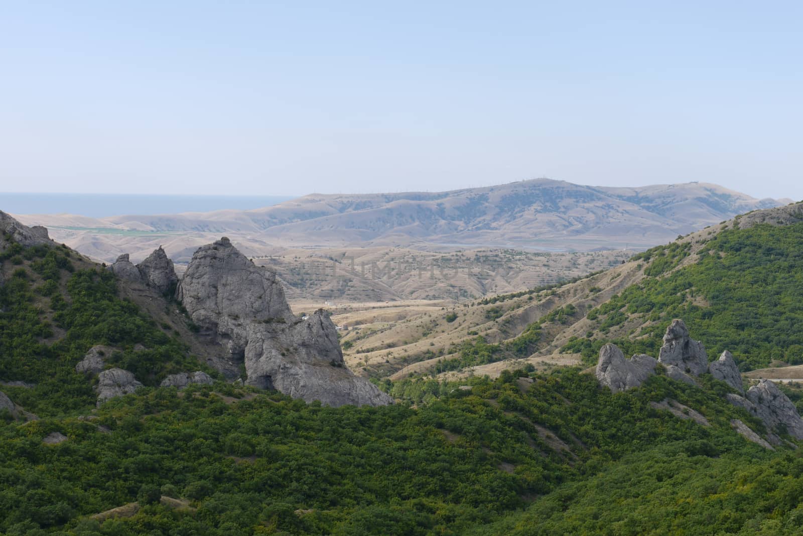 The picturesque nature of the mountainous terrain with slopes overgrown with green lush trees and completely without vegetation with the blue sea visible far away. by Adamchuk