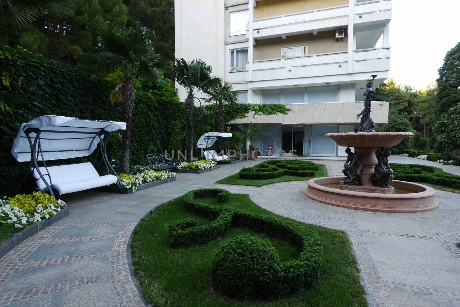 A chic interior courtyard near the hotel with an elegant fountain with sculptures by a designer lawn and white benches with a canopy with beautiful flowers growing next to it.