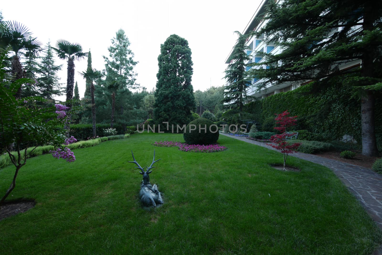 Statue of a deer on a lawn among a flower bed and trees by Adamchuk