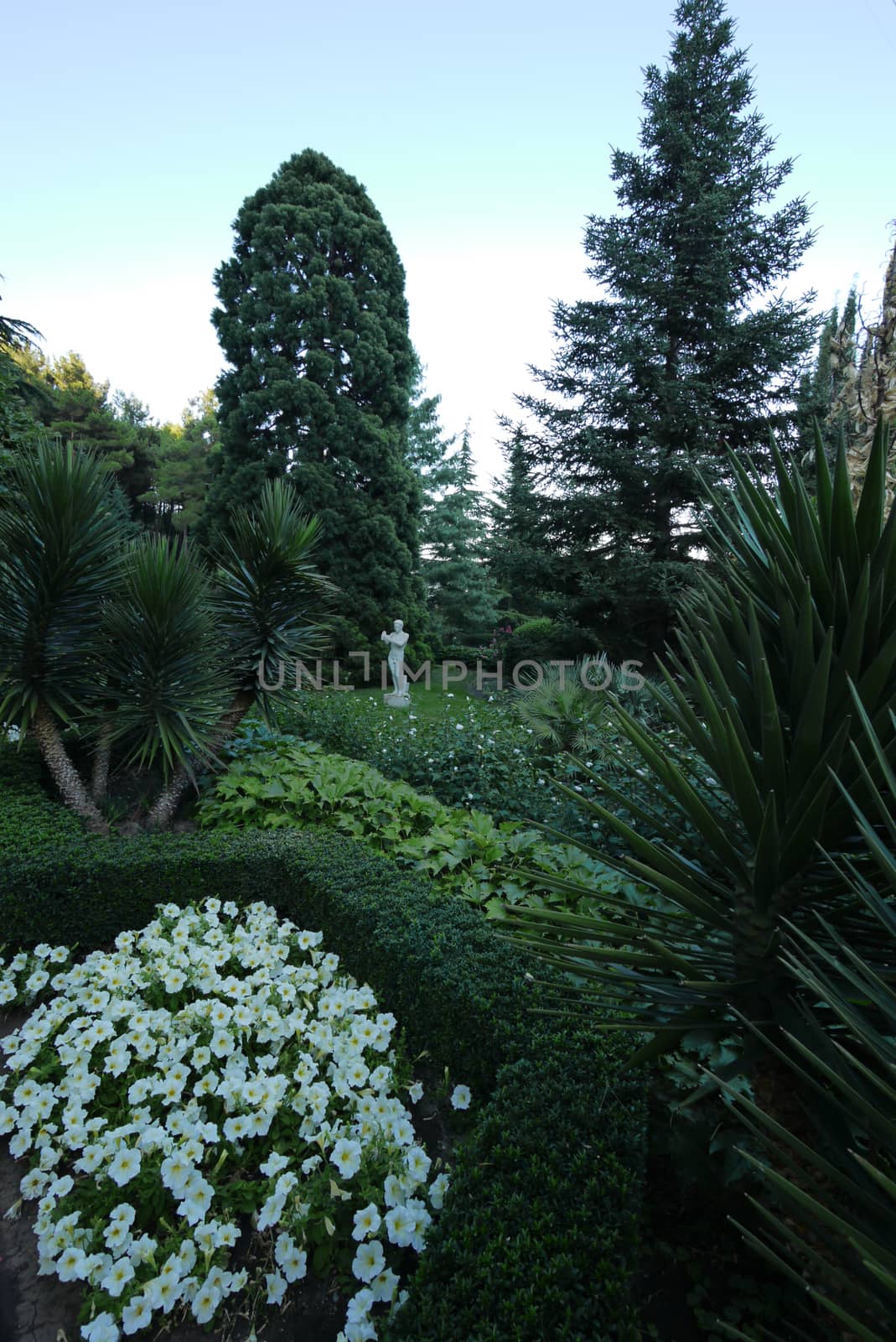 A beautiful white sculpture of gypsum lurking near a green tree next to the lush green flower beds with different vegetation