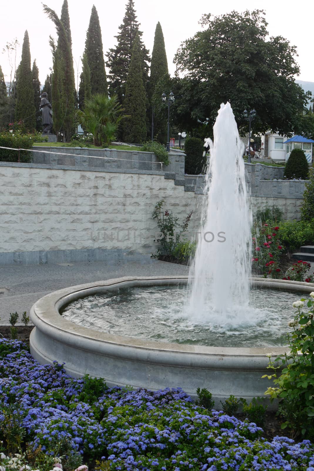 A magnificent fountain with clear water with a beautiful flower bed with flowers on the background of a monument standing in the distance among the tall lush trees.