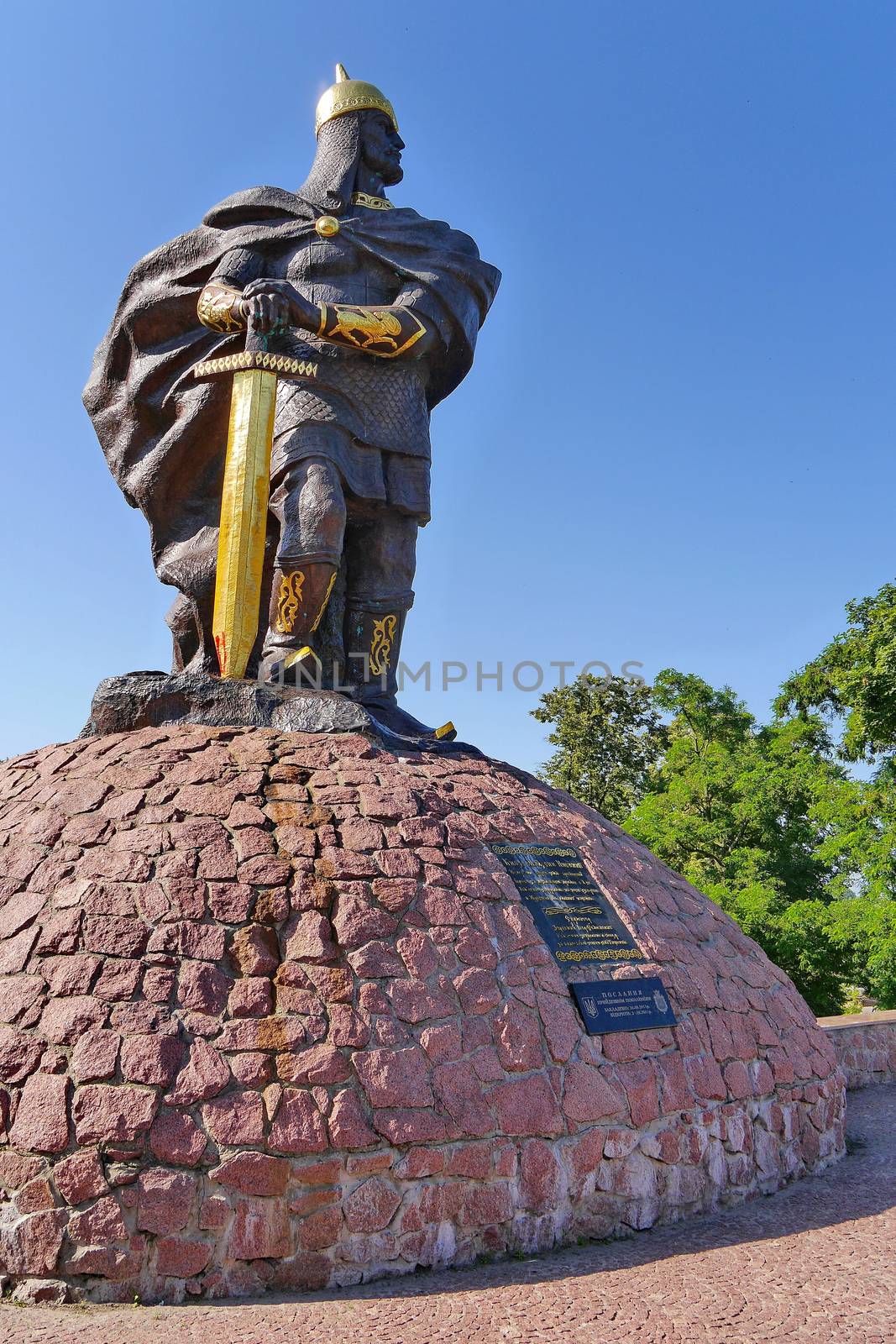 High statue of a knight under a blue sky on the background of beautiful green trees by Adamchuk