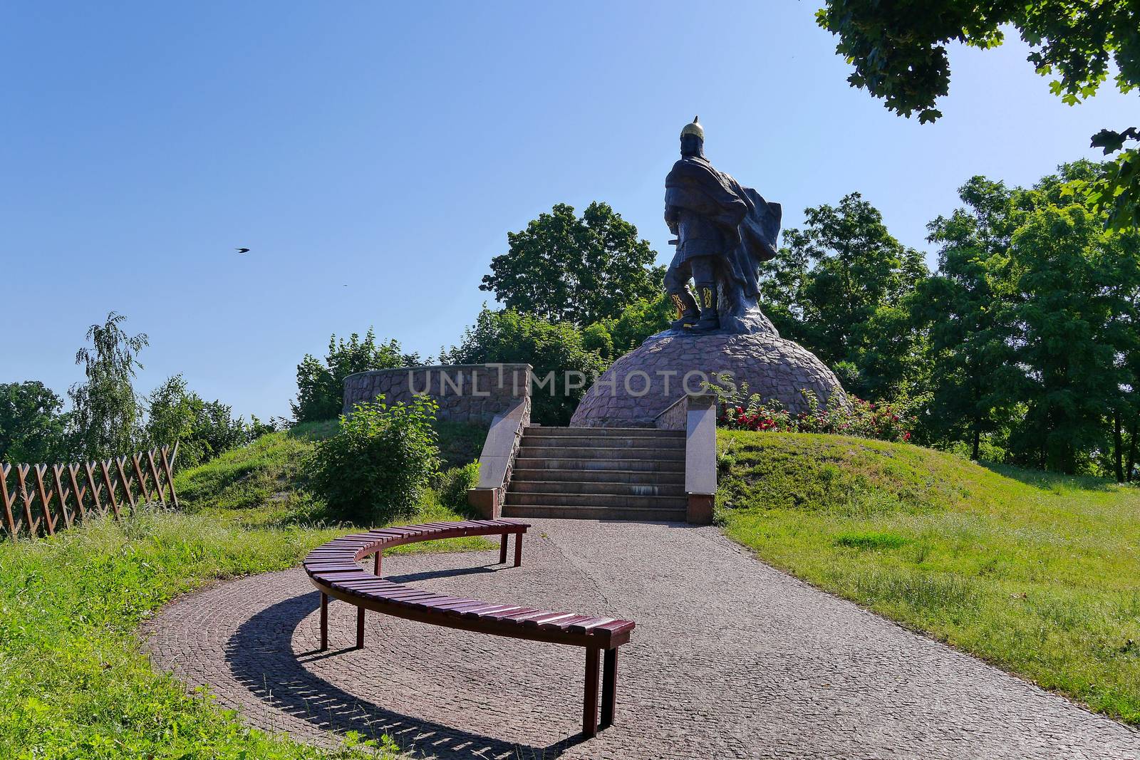 High statue of a knight under a blue sky on the background of beautiful green trees by Adamchuk