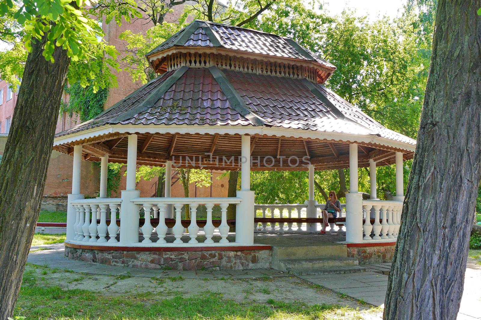 A cozy arbor is surrounded by tall green trees on the background of a tall brick building by Adamchuk