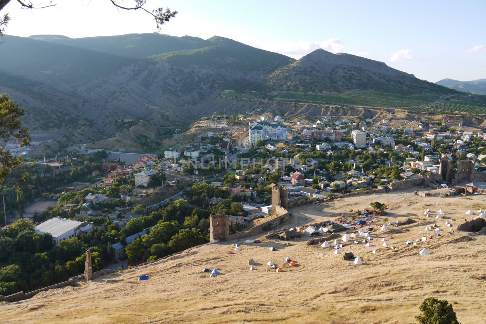 a small cozy town surrounded by high grassy slopes on a blue sky background by Adamchuk