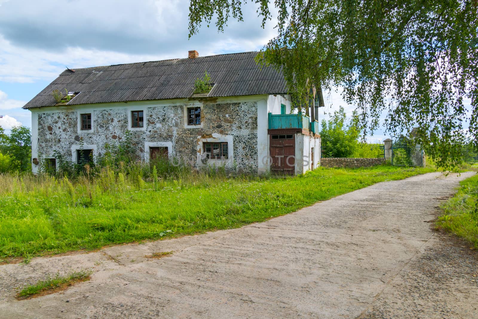 A long wide road is surrounded by grass on the background of abandoned houses and tall, lush trees