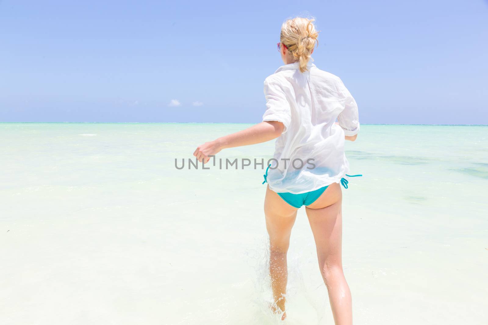 Young slim fit woman wearing white beach tunic running in sea water making water splashes with her legs. Vacation concept. Summer mood. Tropical beach setting. Paje, Zanzibar, Tanzania.