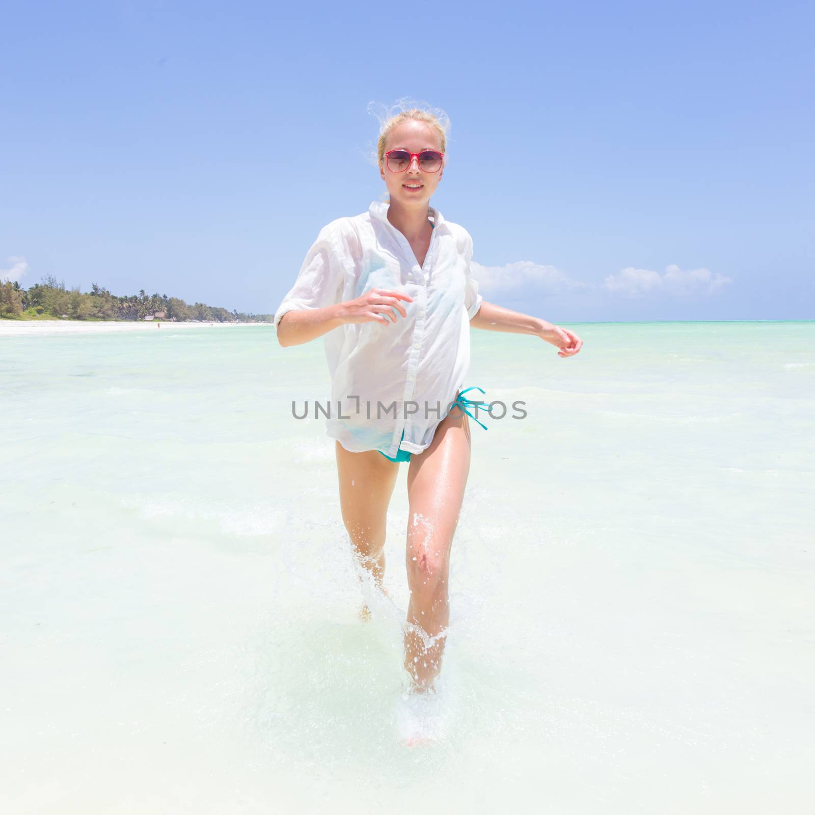 Young slim fit woman wearing white beach tunic running in sea water making water splashes with her legs. Vacation concept. Summer mood. Tropical beach setting. Paje, Zanzibar, Tanzania.