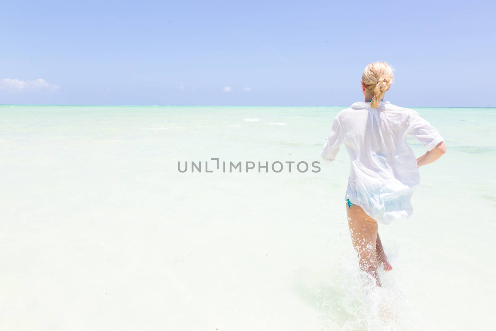 Young slim fit woman wearing white beach tunic running in sea water making water splashes with her legs. Vacation concept. Summer mood. Tropical beach setting. Paje, Zanzibar, Tanzania.