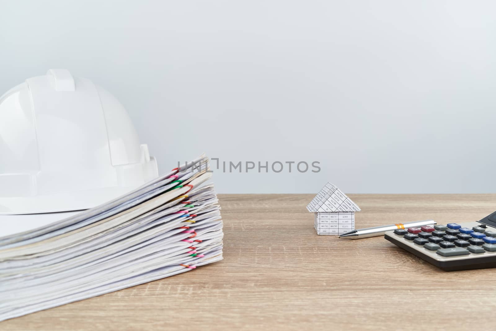 House and pen with calculator have blur white engineer hat on pile overload document of report and receipt with colorful paperclip on wooden table with white background and copy space.