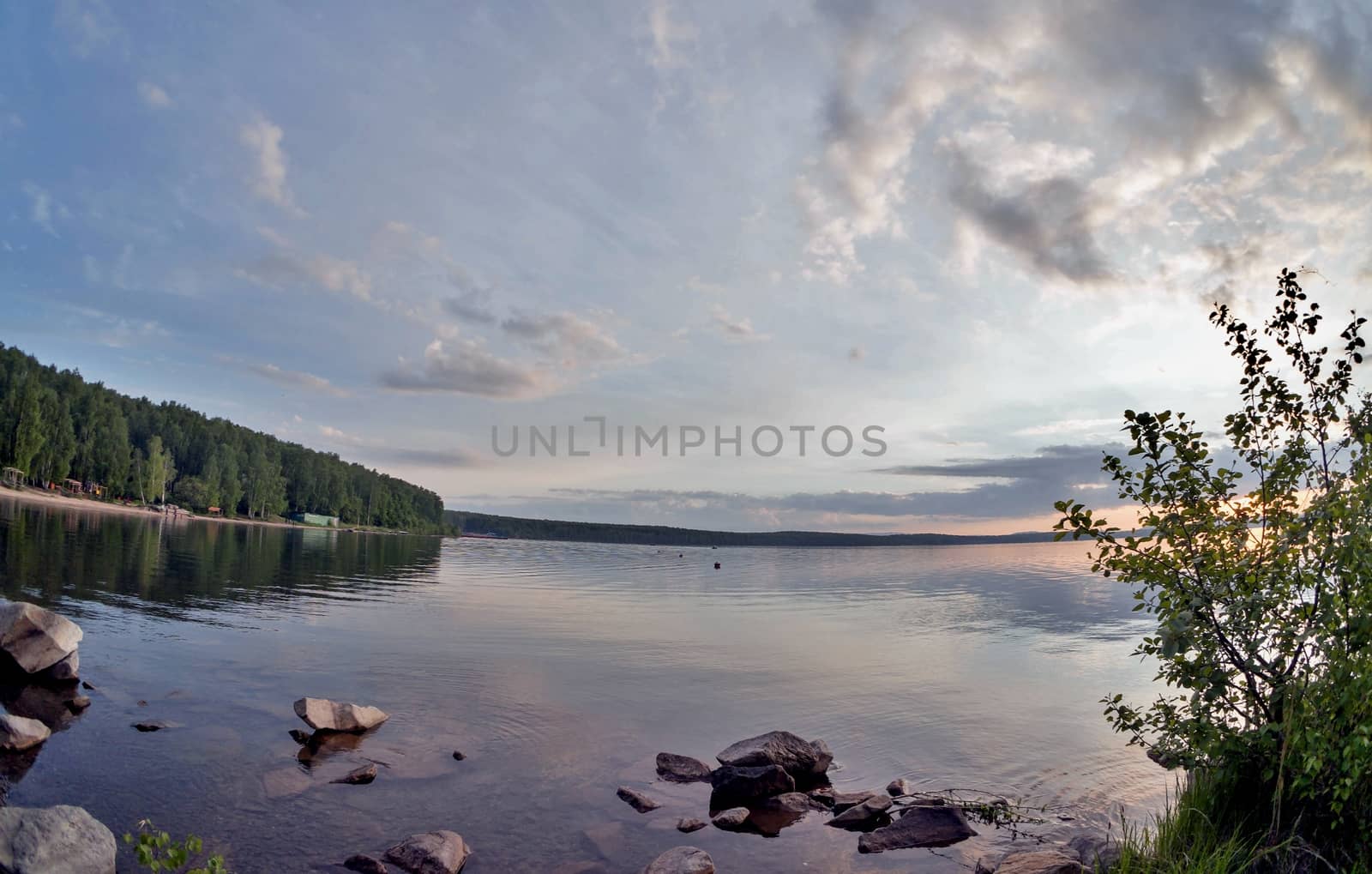 the evening sky over the lake, silhouettes of clouds illuminated by the sun, forest-covered, rocky shore of the lake
