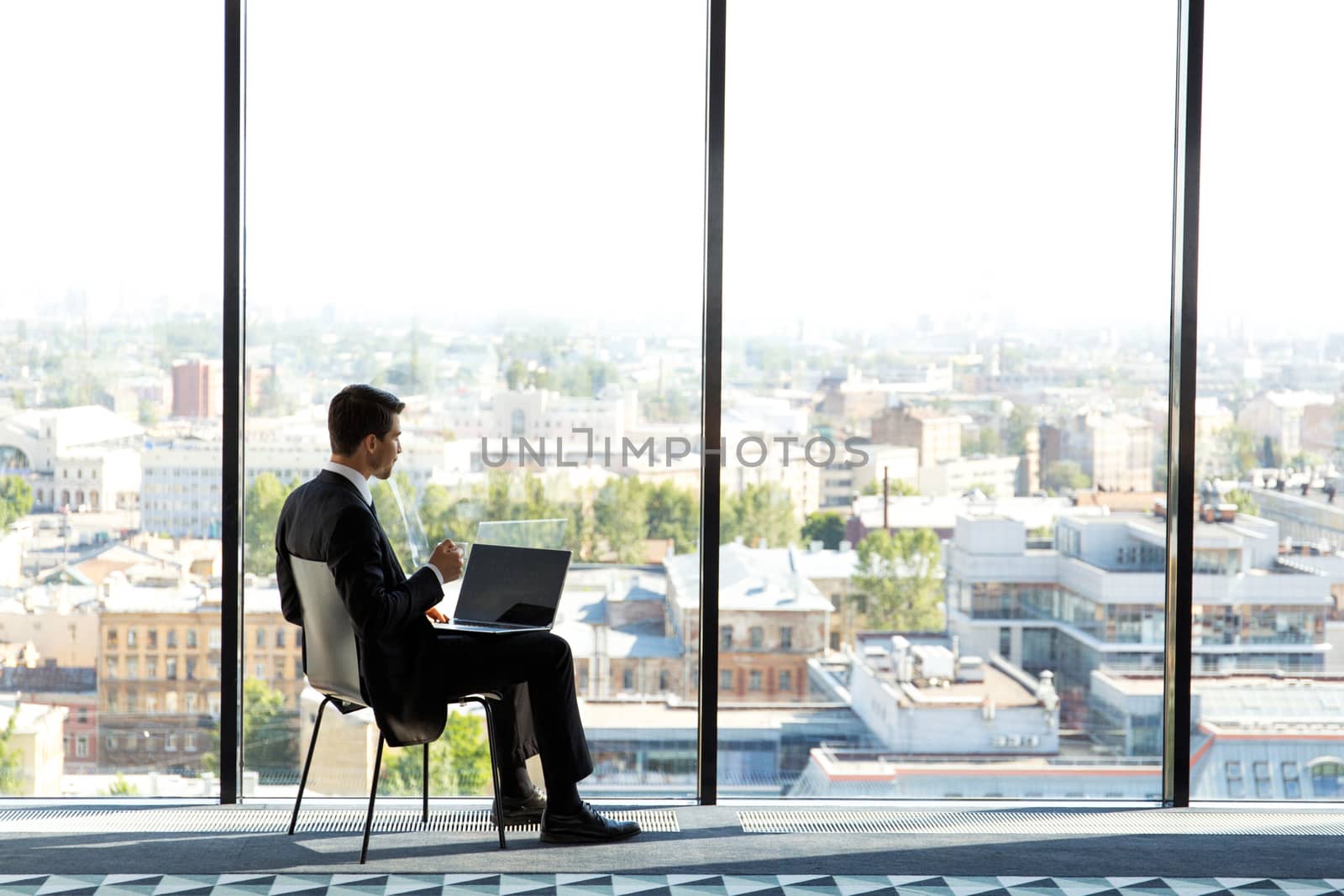 Business man sitting in modern office with panoramic windows, typing on his laptop and srinking coffee