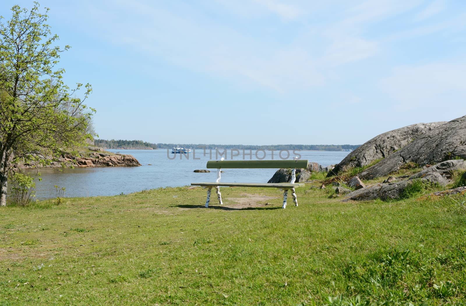 Empty green and white bench on a grassy hilltop above the water on Suomenlinna, a ferry sails past in the background
