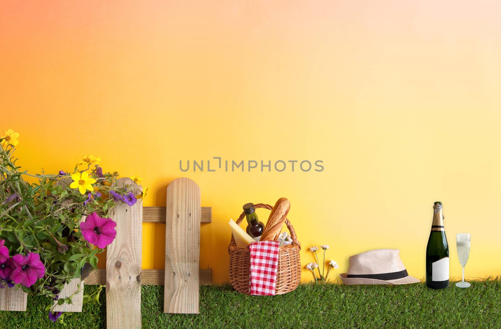 Summer picnic basket with bread, cheese and wine on grass with garden fence and flowers