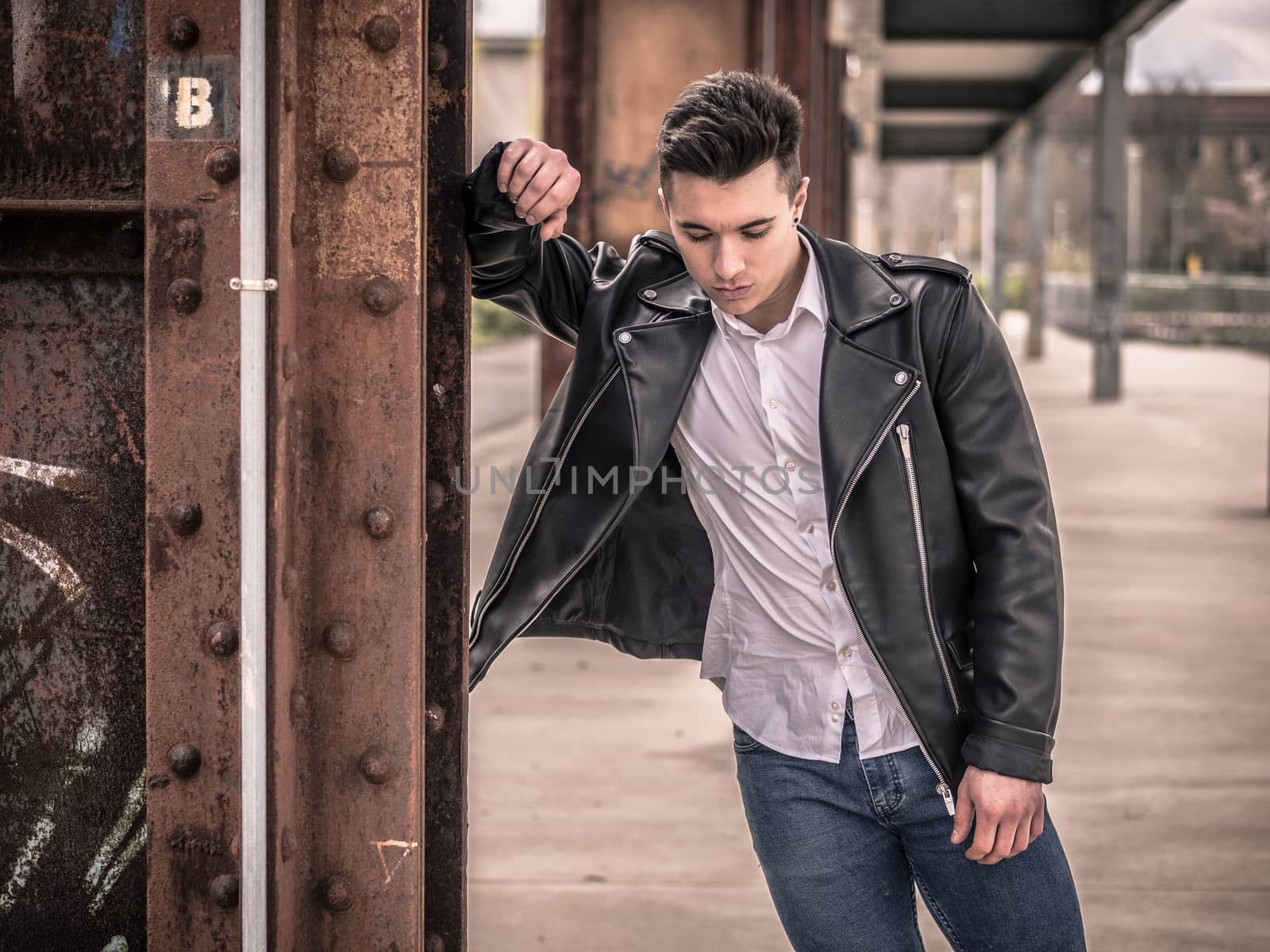One handsome young man in urban setting in European city, standing, wearing black leather jacket and jeans