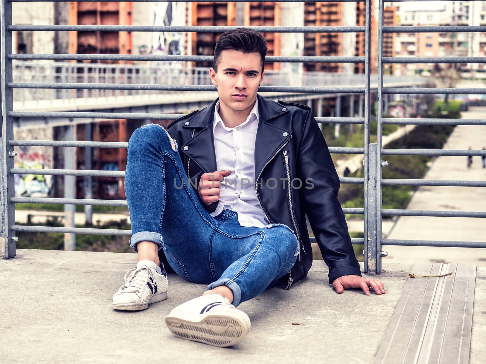 One handsome young man in urban setting in European city, sitting, wearing black leather jacket and jeans