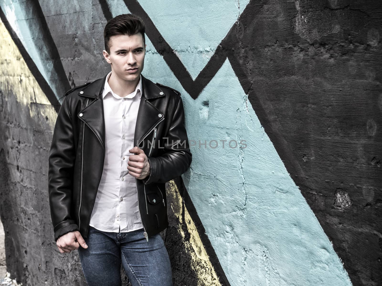 One handsome young man in urban setting in European city, standing, wearing black leather jacket and jeans