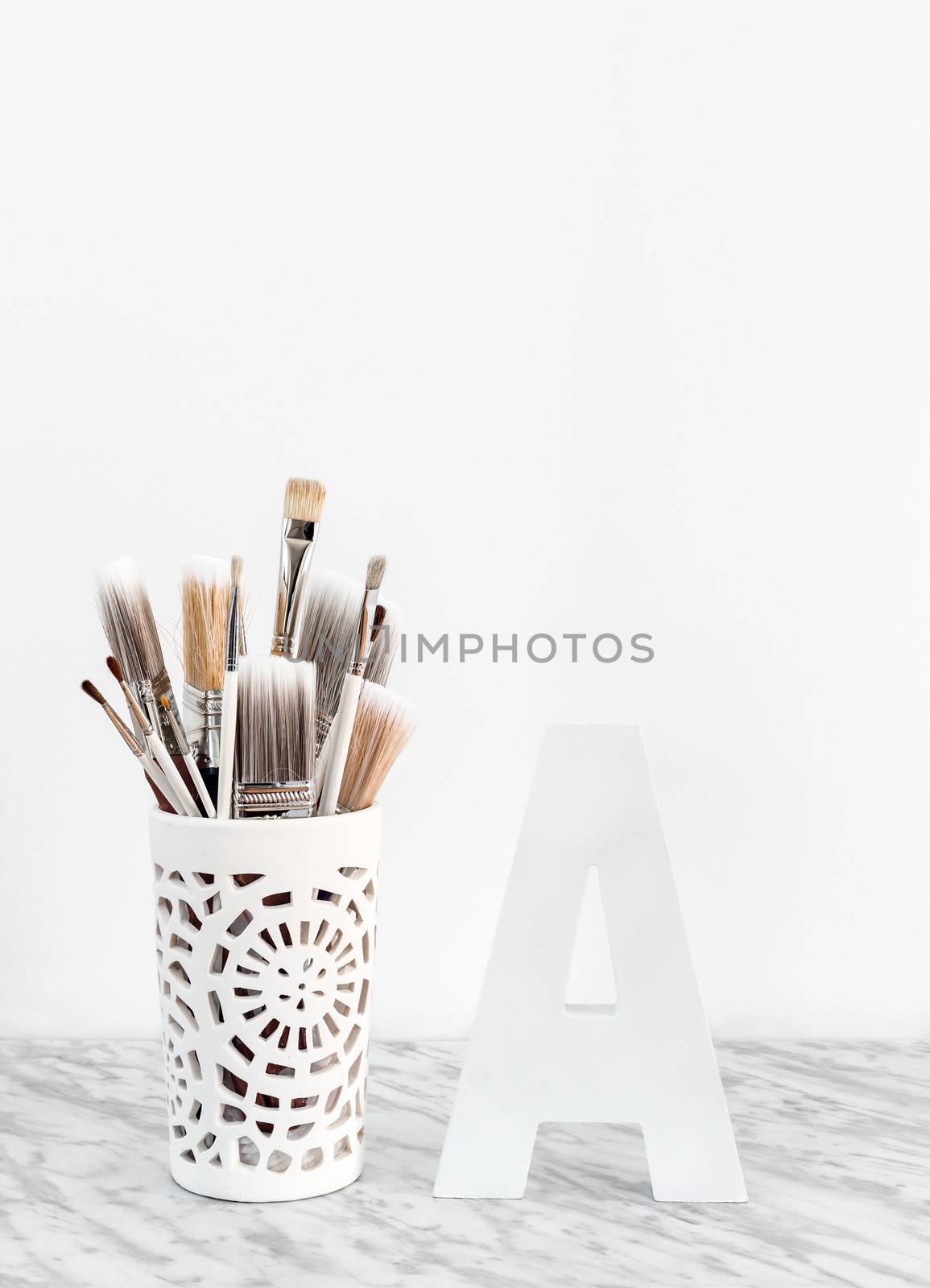 Paintbrushes in a decorative vase and white letter A on marble surface.