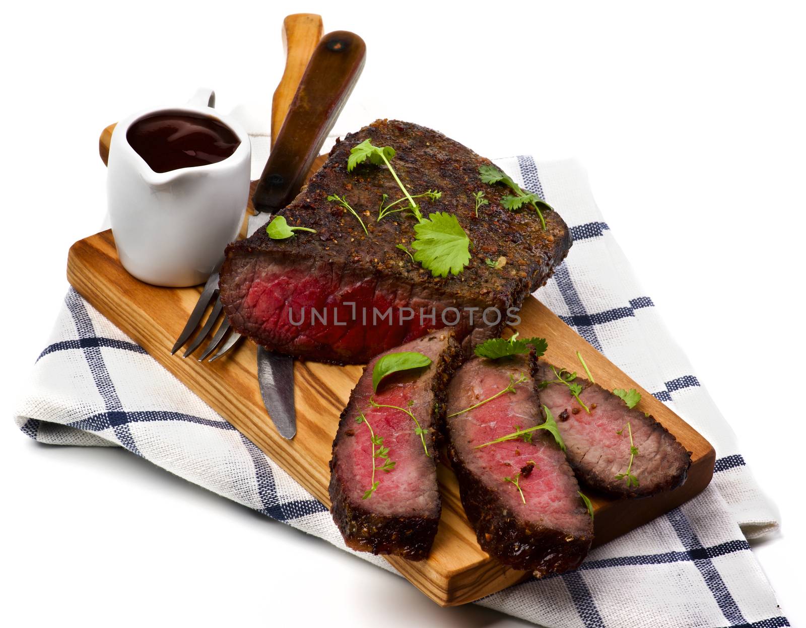 Delicious Roast Beef Medium Rare Sliced on Wooden Cutting Board with Tomato Sauce, Fork and Table Knife on Checkered Napkin closeup on White background