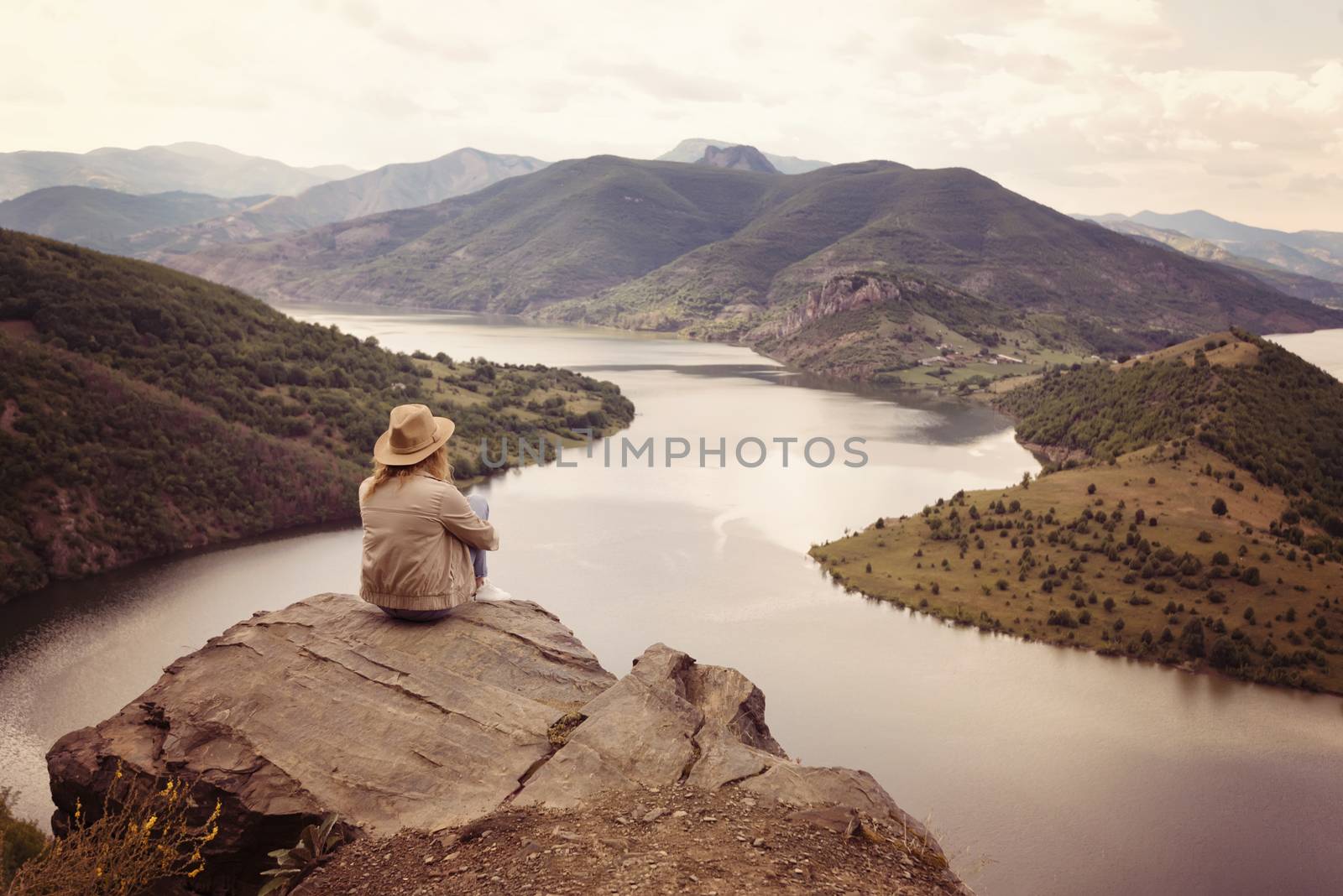 A hiking girl sits on the edge of the cliff and looking at the river and mountains. Location: the meanders of Arda river, Bulgaria