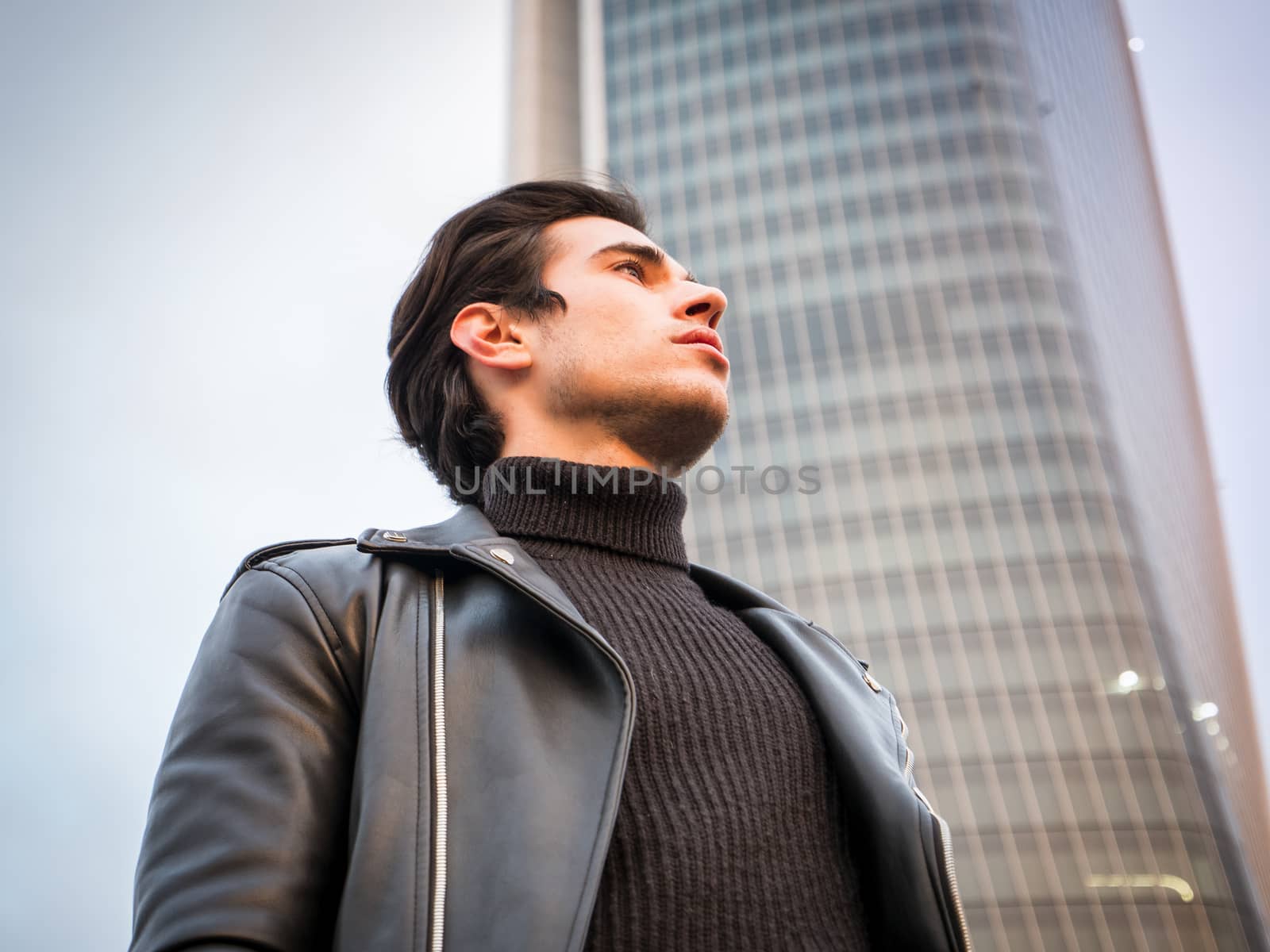 One handsome young man in urban setting in modern city, standing, wearing black leather jacket and jeans